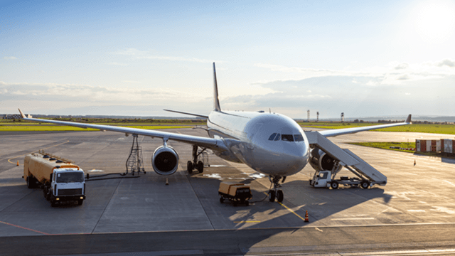 Commercial airplane parked at airport gate with boarding stairs, fuel truck, and ground support vehicles during sunset